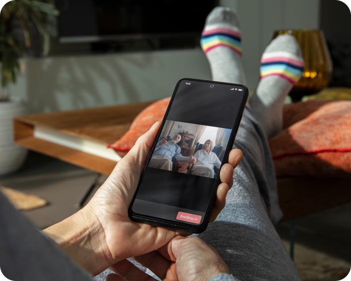 A person with their feet up on their coffee table is using the JubileeTV app on their cell phone to checking on their parents; who are enjoying an evening of television while holding hands.