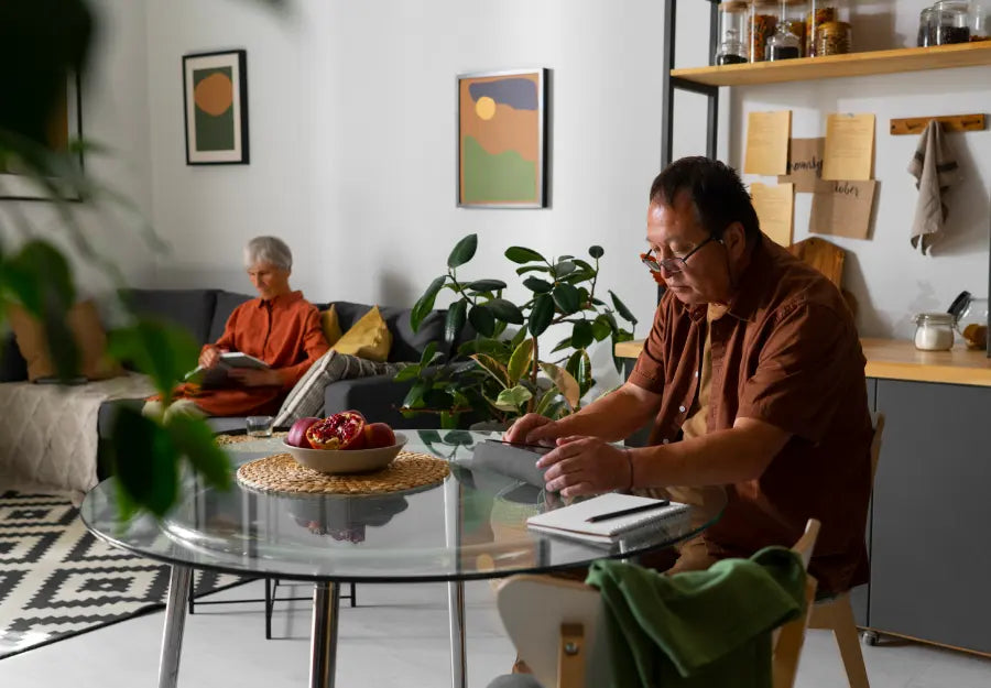 A caregiver is sitting at the table with a notebook while an elderly woman reads a book in the background