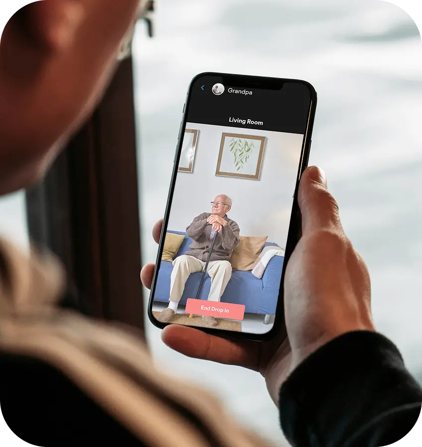 Person holding a smartphone with a one-way remote monitoring video call to an elderly person on a couch.