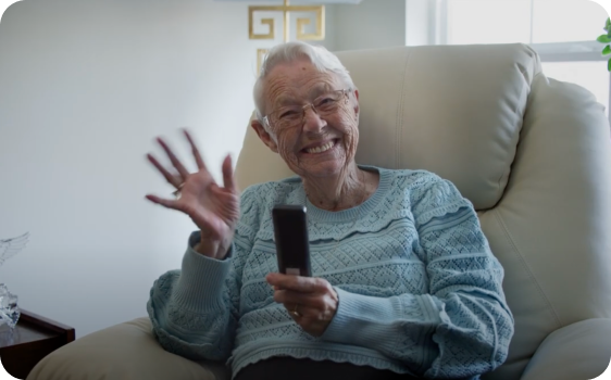 An elderly woman siting in a recliner smiling and waving at the camera with a remote in her other hand.