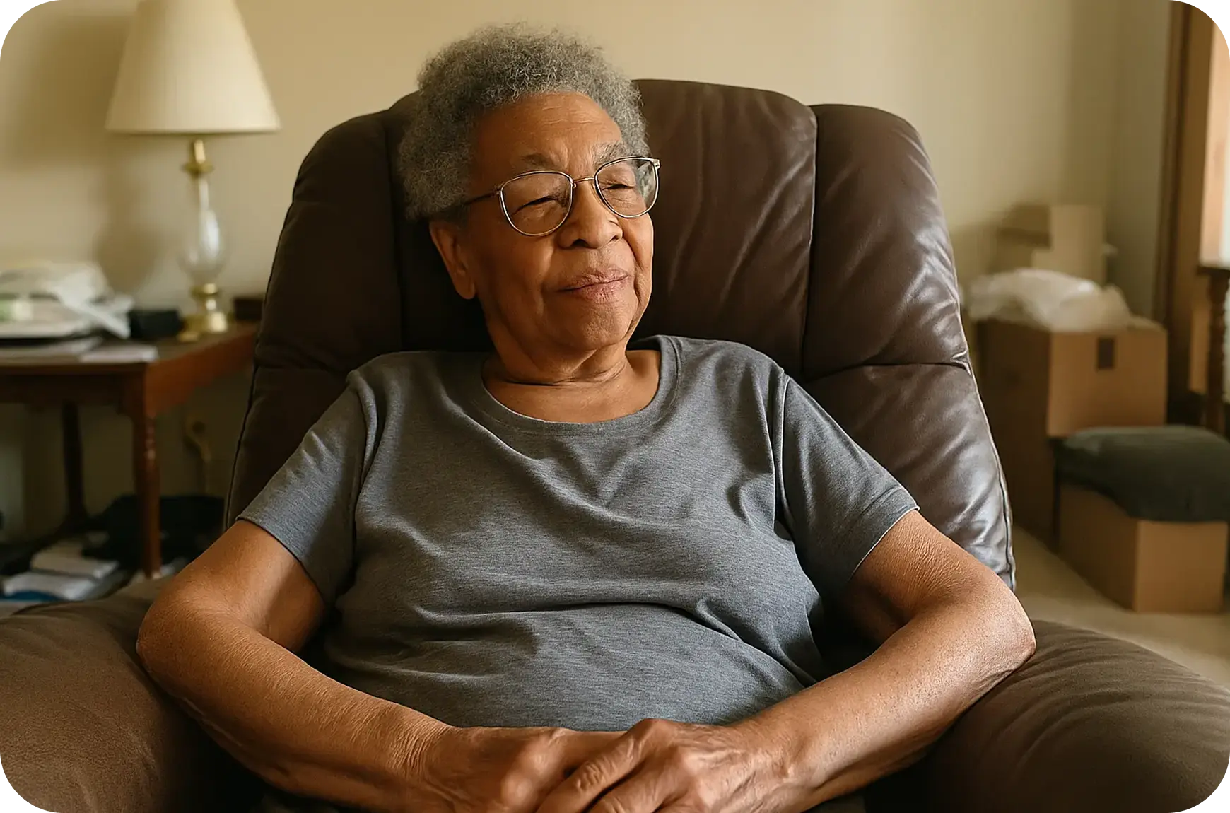 An elderly woman sitting in a brown armchair wearing glasses, with her hands in her lap looking content with a slight smile as she looks off to the left.