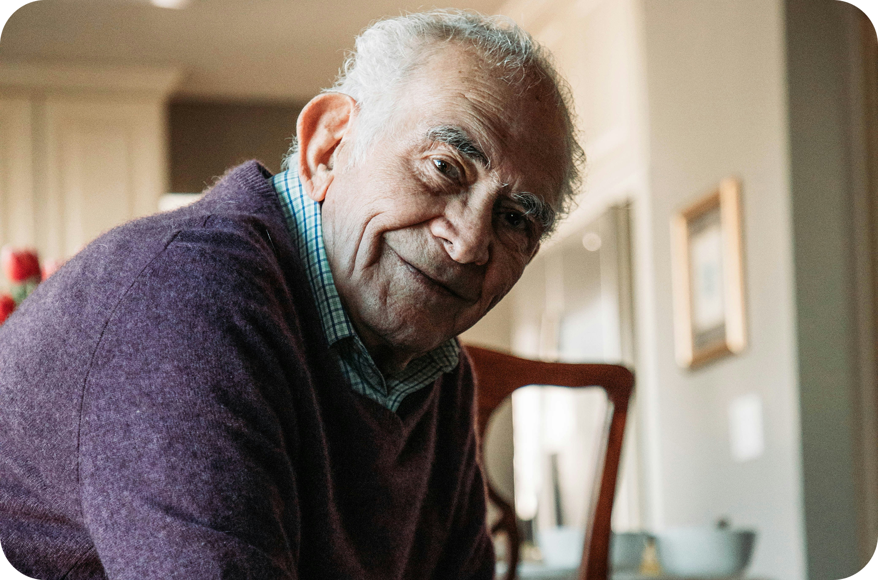 Man in a purple sweater sitting in a home setting smiling lovingly at the person behind the camera.