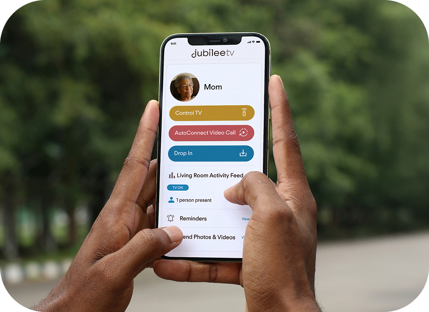 Person holding a smartphone displaying the Jubilee TV app with a blurred green outdoor background.  They are checking in on their loved one and can see that they are alone and the living room TV is on.