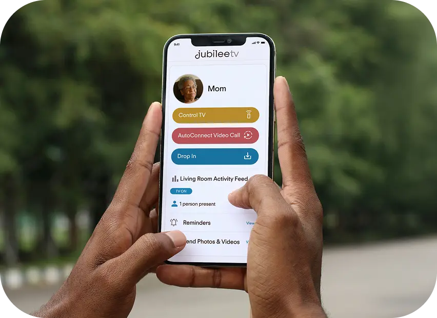 Person holding a smartphone displaying the Jubilee TV app with a blurred green outdoor background.  They are checking in on their loved one and can see that they are alone and the living room TV is on.