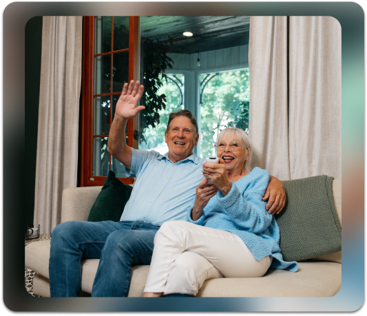 elderly couple sitting on couch waving to video call on TV