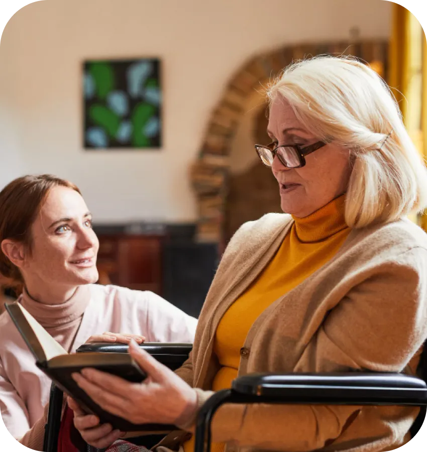 Two women sitting together, one holding a book, in an assisted living community.
