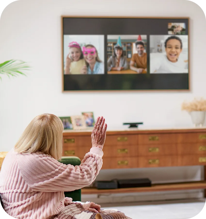 Elderly woman sitting in front of a television screen displaying a video call with four people.