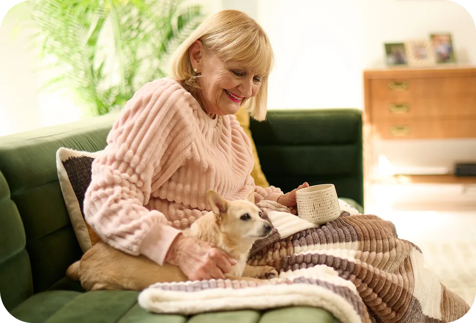 Woman sitting on a couch with a small dog, holding a mug, in a cozy living room setting.