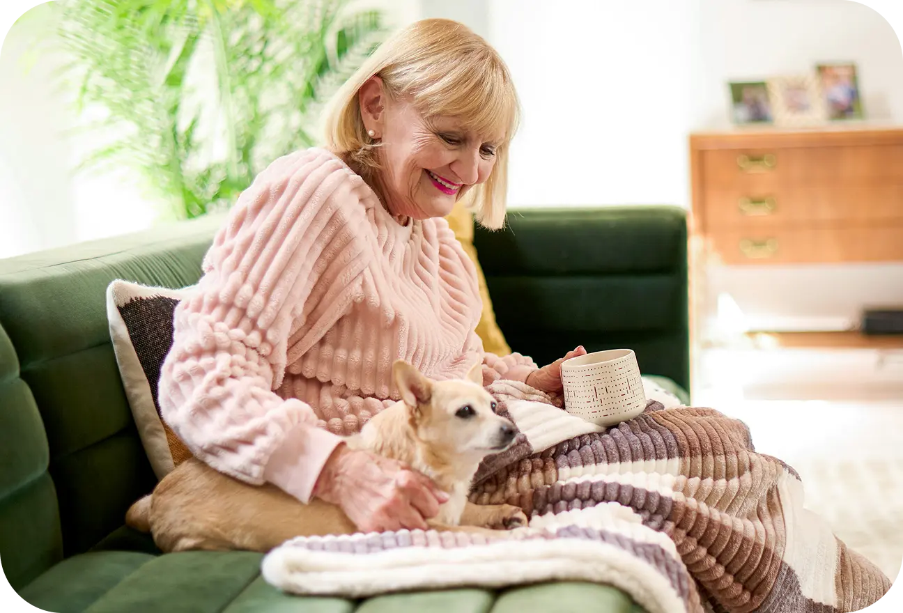 An elderly woman enjoying a hot beverage and a snuggle with her dog on the couch with a fuzzy blanket.